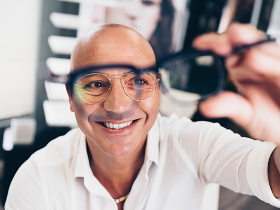 Bald man holding up a pair of glasses looking through one of the lenses