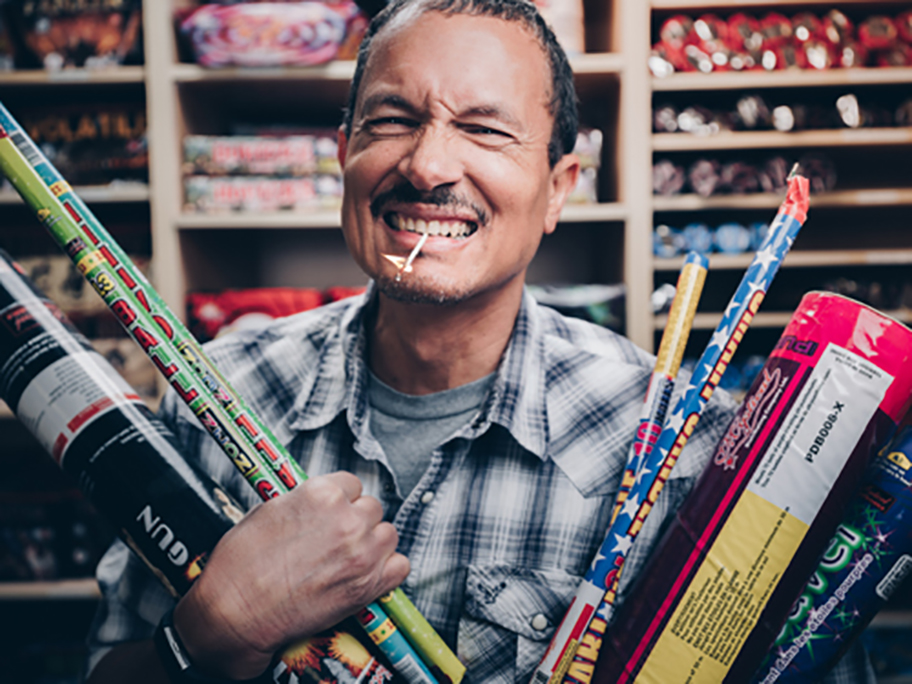 A man holding a bunch of fireworks in both arms with a lit match in his mouth