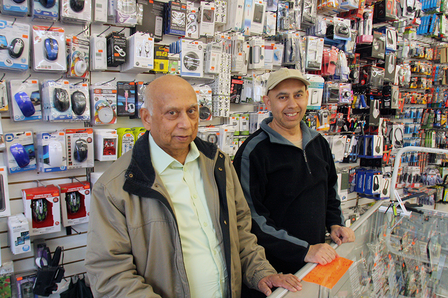 Photo of two men standing behind the counter at the dollar store they own. The wall if full of gadgets and items for sale.