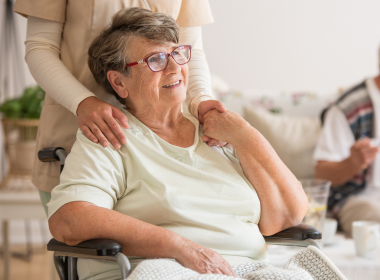 A photo of an older person in a wheelchair holding the hand of a nurse standing behind them.