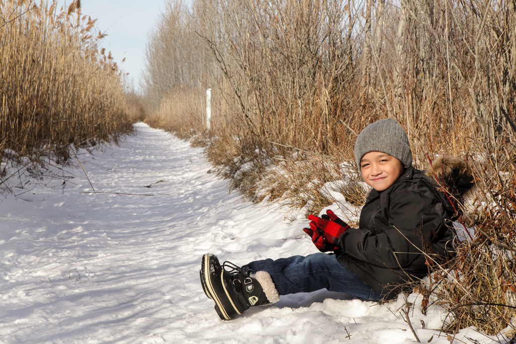 A photo of a young child sitting in the snow at the side of a trail.
