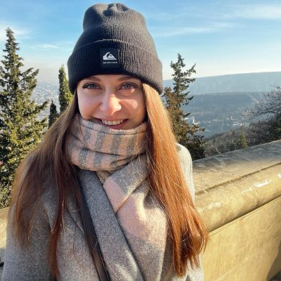 A photo of a young woman with long hair wearing a black toque standing at a lookout overlooking a city staring into the camera.