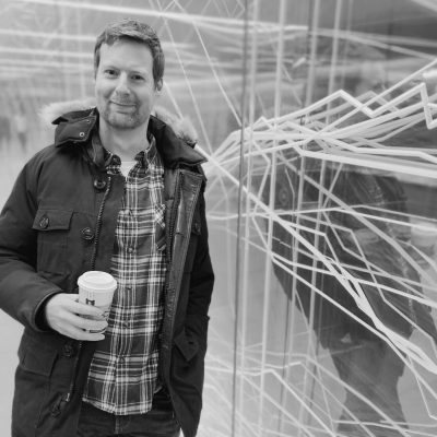 A black and white photo of a person in a winter jacket holding a to go cup of coffee standing in front of a wall with squiggly lines on it.